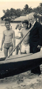 An old black and white picture of Ford (in suit) with youths in swim trunks on a beach.