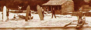 An old sepia photograph of people holding surfboards on the beach. In the background is a grass building and to the right is a wooden outrigger canoe. 