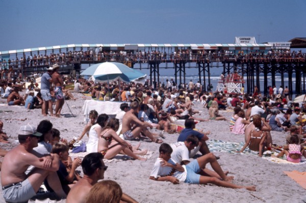An old photograph of a large group of people on the beach with the pier in the background.