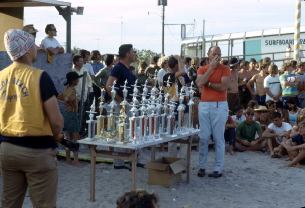 A group of people on the beach gathered around a table with many trophies on it.