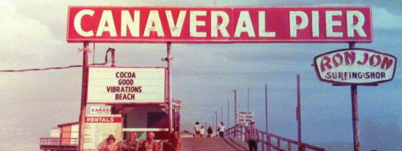 Historical Photo of Canaveral Pier with the pier extending into the distance. At the top, a large red and white sign reads: Canaveral Pier. On the left, a smaller black and white sign reads: COCOA GOOD VIBRATIONS BEACH. On the right is a Ron Jon Surfing Shop sign.